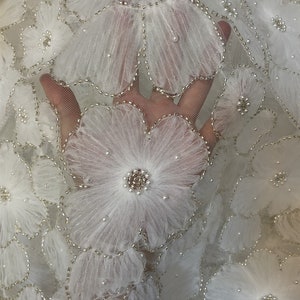 以下が含まれることがあります： 3Dの花とパールがあしらわれた白い花のレース生地。生地は繊細で透き通っており、柔らかく流れるような質感です。