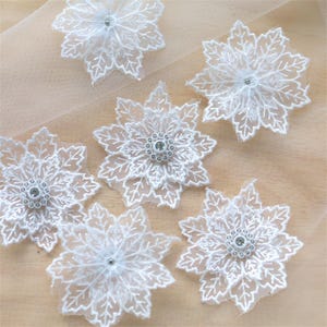 May include: A collection of six white floral lace appliqués, each featuring intricate leaf-like petal embroidery and a central circular embellishment adorned with small clear crystals, arranged on a sheer, light beige fabric background.