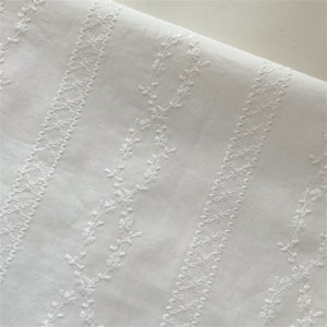 May include: White cotton fabric with a delicate embroidered pattern of repeating floral vines. The embroidery is in a white thread and creates a subtle texture on the fabric.