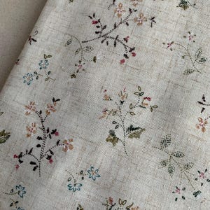May include: A light beige fabric featuring a repeating pattern of small embroidered floral sprigs. The delicate flowers are stitched in shades of blue, pink, brown, and green, creating a textured surface with visible woven linen-like threads.