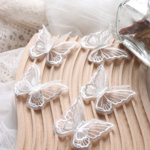 May include: Delicate white butterfly appliques with intricate embroidered details. The butterflies are arranged on a light wooden surface, with a soft, sheer fabric in the background. These decorative appliques are ideal for craft projects and embellishments.