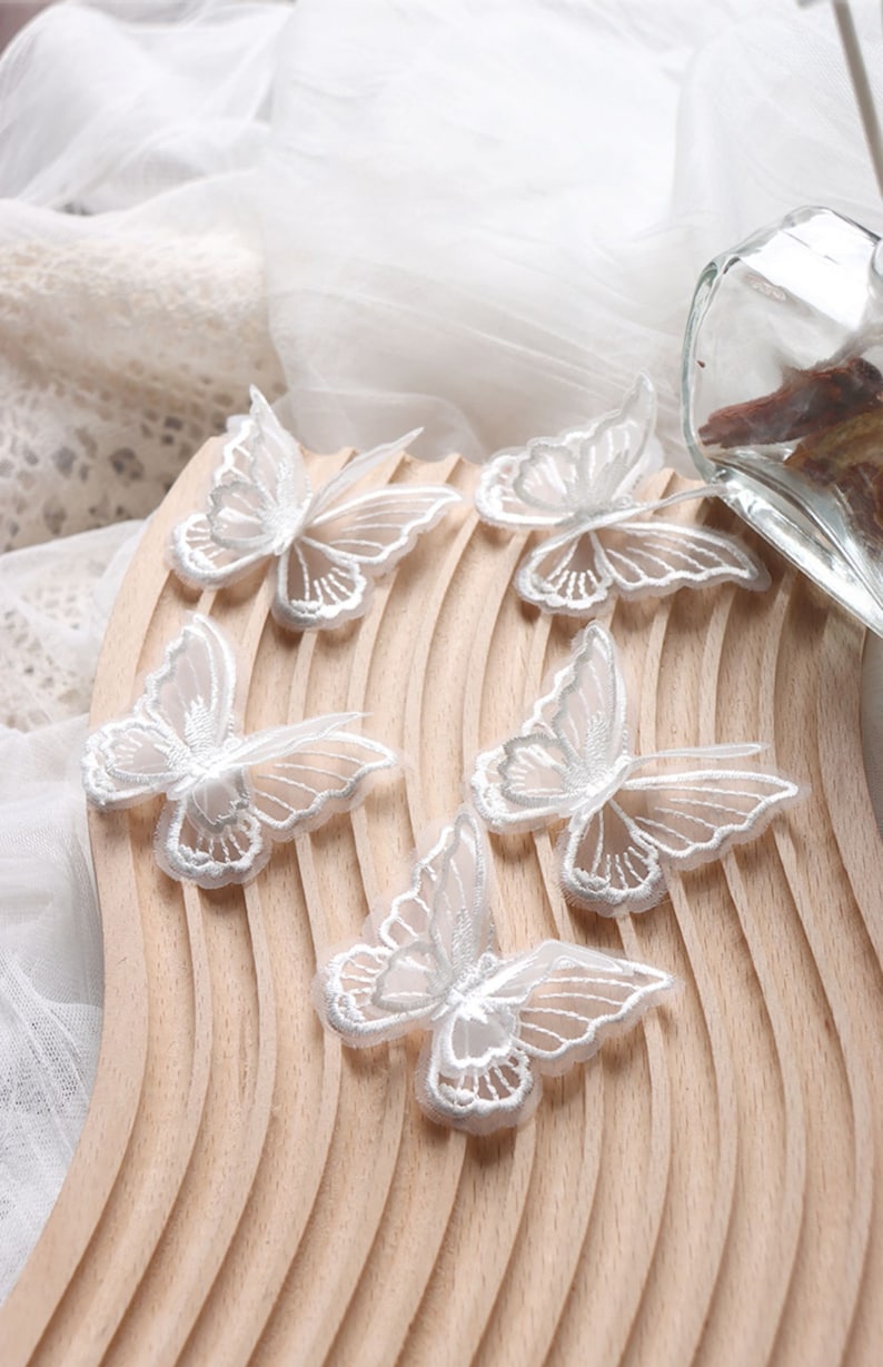May include: Delicate white butterfly appliques with intricate embroidered details. The butterflies are arranged on a light wooden surface, with a soft, sheer fabric in the background. These decorative appliques are ideal for craft projects and embellishments.