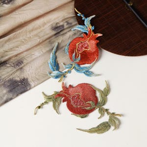 May include: Two embroidered iron-on patches featuring red pomegranates with green and blue leaves.
