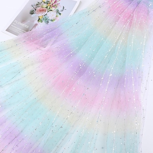 Könnte beinhalten: Ein durchscheinender, schillernder Stoff mit einem Regenbogenverlauf aus Farben, darunter Rosa, Lila, Blau und Gelb. Der Stoff ist mit kleinen, schimmernden silbernen Punkten besetzt. Das Wort "FLOWERS" ist auf einer Zeitschrift im Hintergrund zu sehen.