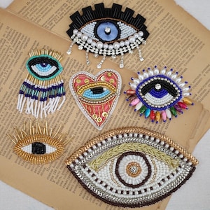 May include: A collection of five embroidered patches featuring eye and heart motifs. The designs are crafted with beads, sequins, and metallic threads in various colors including blue, gold, white, black, and pink, showcasing intricate textures.