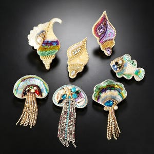 May include: A collection of seven decorative brooches featuring marine-themed designs including seashells, a fish, and jellyfish. These accessories are crafted with colorful sequins, beads, crystals, and dangling rhinestone fringe accents.