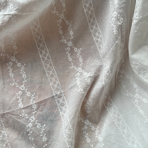 May include: White cotton fabric with delicate embroidered floral patterns in a vertical stripe design.