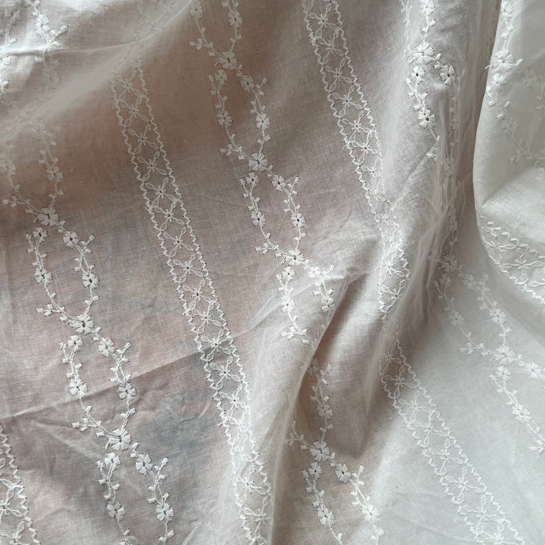 May include: White cotton fabric with delicate embroidered floral patterns in a vertical stripe design.