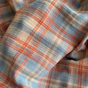 May include: A lightweight fabric featuring a plaid pattern with intersecting lines in shades of orange, light blue, and cream. The material has a visible crinkled texture throughout, creating a soft, layered appearance with folds and depth.