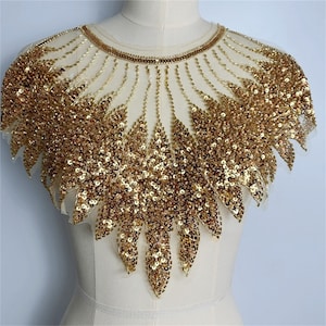 Könnte beinhalten: Goldene Pailletten-Applikation mit floralem Design. Die Applikation besteht aus goldenen Pailletten und Perlen, die auf einem transparenten, cremefarbenen Netzstoff aufgenäht sind. Das Design ist symmetrisch.