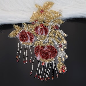 May include: An embroidered floral applique with gold, silver, and red beadwork. The design features three red flowers with beaded fringe, gold leaves, and silver accents. The applique is on a sheer mesh backing.