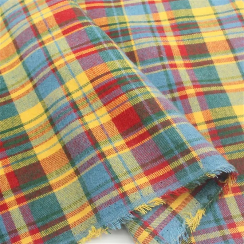 K&ouml;nnte beinhalten: Nahaufnahme eines farbenfrohen karierten Stoffes mit ausgefranstem Rand. Das Muster zeigt eine Mischung aus roten, gelben, blauen und gr&uuml;nen sich kreuzenden Linien, die ein klassisches Tartan-Design bilden. Der Stoff wirkt weich und strukturiert.