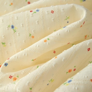May include: Close-up of a cream fabric with a delicate floral pattern. The fabric has a textured surface with small, raised dots and vertical striped details. Tiny flowers in red, blue, orange, and green are scattered across the material.