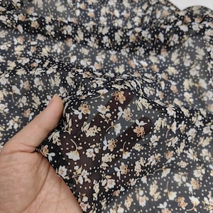 Puede incluir: Una tela negra transparente con un patrón denso de pequeñas flores blancas y beige intercaladas con diminutos puntos blancos. El material ligero es semitransparente, revelando la textura y el color de la superficie que hay debajo.