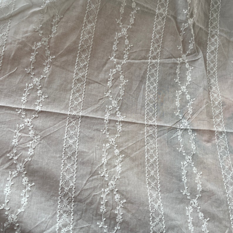 May include: White cotton fabric with a delicate embroidered pattern of small flowers and vines. The fabric has a sheer, lightweight texture.