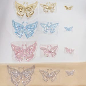 May include: A collection of twelve butterfly-shaped appliques arranged in four rows by color, including gold, light blue, pink, and silver. Each row features three sizes of butterflies decorated with sequins and beads on a sheer mesh backing.