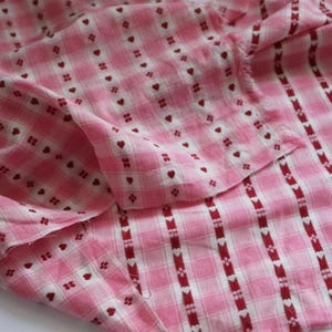 Pode incluir: Tecido xadrez rosa e branco com padrão de coração. O tecido apresenta um padrão repetitivo de pequenos corações vermelhos e detalhes florais. O tecido está dobrado, mostrando a textura e o design.