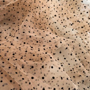 Può includere: Primo piano di un tessuto beige trasparente con un motivo a pois neri. Il tessuto è drappeggiato, creando morbide pieghe e ombre. I pois neri sono distribuiti uniformemente sul materiale, aggiungendo un tocco giocoso alla texture elegante.