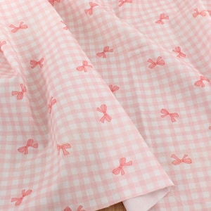 Può includere: Tessuto a quadretti rosa e bianco con un motivo ripetuto di piccoli fiocchi rosa. Il tessuto è piegato, mostrando la trama e i dettagli del motivo. L'insieme è morbido e femminile, adatto a vari progetti creativi.