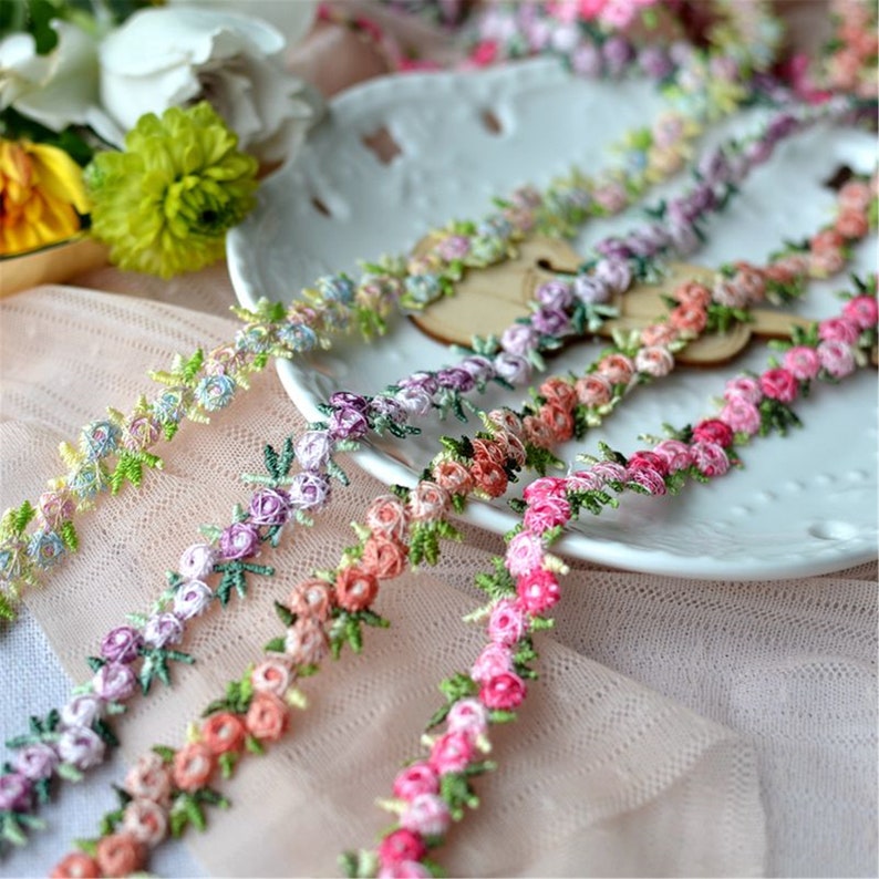 May include: Assortment of delicate floral lace trim in various pastel colors, featuring small embroidered flowers and leaves. The lace is arranged on a white plate and a pink fabric background.