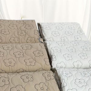 May include: A set of quilted bedding covers featuring a repeating flower pattern. The collection includes beige and white fabric options, both decorated with dark outlines of five-petaled flowers stitched into the soft, textured material.