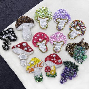 May include: A collection of eleven embroidered patches featuring various mushroom and fruit designs. The patches are crafted with colorful glass beads and small white pearls, depicting mushrooms in shades of brown, green, purple, red, pink, and yellow, alongside two clusters of grapes in green and purple.