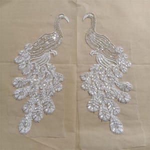 Può includere: Due applicazioni simmetriche a forma di pavone, realizzate con filo bianco e argento e paillettes riflettenti. Ogni figura di uccello presenta un collo lungo, una testa curva e un elaborato design a coda a ventaglio, ricamato su rete.
