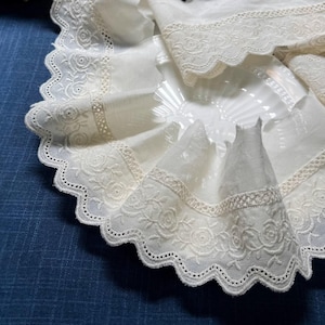 Può includere: Bordura in pizzo di cotone bianco con bordo smerlato e delicato ricamo floreale. Il pizzo è mostrato piegato su un piatto bianco.