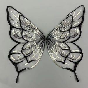 Può includere: Un paio di ali di farfalla simmetriche con contorno nero e dettagli ricamati bianchi e argento. Le ali hanno un design delicato e a merletto, ideale per costumi o progetti artigianali. Larghezza di circa 30 cm.