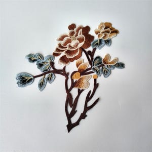 Pode incluir: Aplique floral bordado com flores castanhas, bege e douradas, e folhas azul-esverdeadas. O design inclui um caule e ramos castanhos escuros, sobre um fundo branco. Este patch decorativo é adequado para trabalhos de costura.