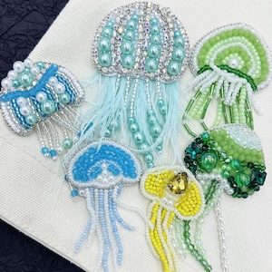 Op de afbeelding: Een collectie van zes handgemaakte kwallenbroches met ingewikkeld kralenwerk, pailletten en kristallen. De ontwerpen bevatten diverse tinten blauw, groen en geel, met bungelende kralententakels en zachte veren.