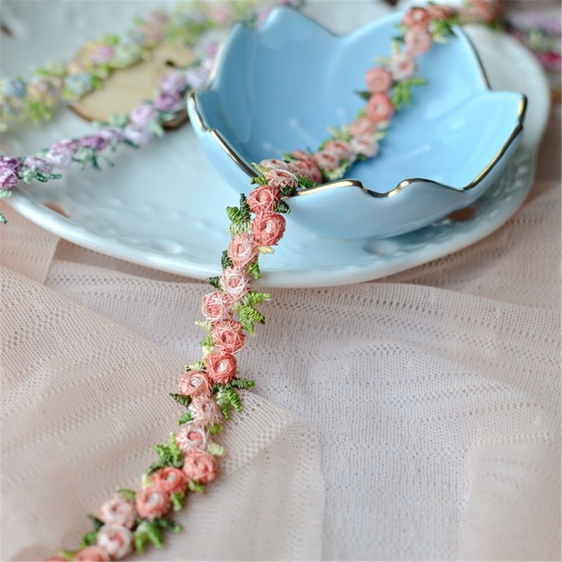 May include: A delicate pink and green floral trim with small, clustered flowers. The trim is displayed on a light blue saucer and a light pink mesh surface.