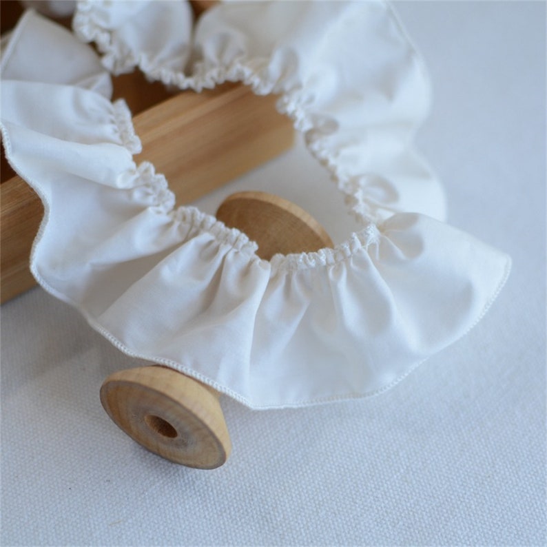 May include: Close-up of a cream-colored, ruffled fabric trim. The trim is gathered and sewn, creating a wavy edge. A small, wooden object with a circular base and a hole in the center is in the foreground. A light-colored wooden box is in the background.