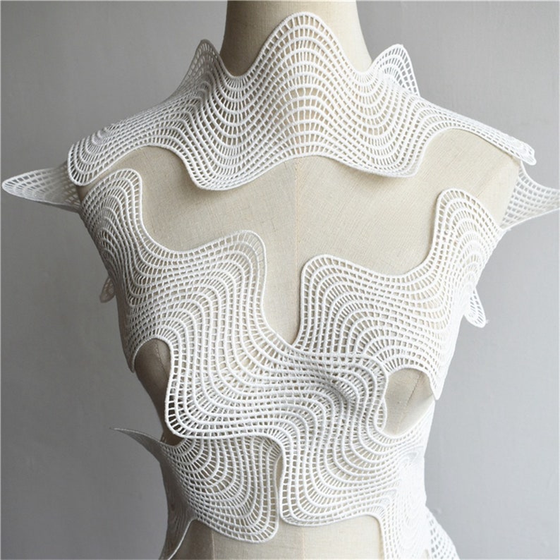 May include: White crocheted fabric with a wavy, geometric pattern. The fabric is draped over a dress form.