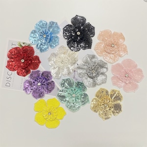 Może przedstawiać: Zestaw dziesięciu aplikacji kwiatowych z cekinami w różnych kolorach, w tym czerwony, niebieski, fioletowy, żółty, zielony, różowy, biały, srebrny, złoty i czarny. Każda aplikacja ma centralny kryształ.