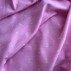 Op de afbeelding: Roze stof met een herhaald patroon van witte strikken en harten. De stof is gevouwen, waardoor schaduwen en textuur ontstaan. De strikken zijn gelijkmatig verdeeld en uitgelijnd met verticale stippellijnen.