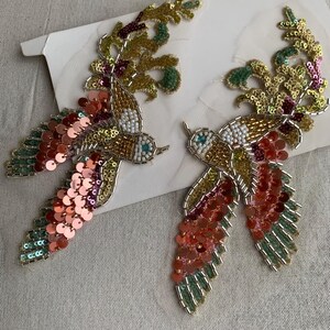 May include: A pair of bird-shaped patches featuring intricate beadwork and sequins in shades of gold, copper, pink, and green. The designs include white beaded accents, metallic gold thread detailing, and layered circular sequins forming the wings and tail feathers.