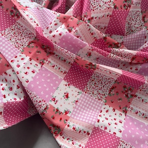 Könnte beinhalten: Ein Patchwork-Stoff mit verschiedenen rosa- und weißfarbenen Mustern. Das Design umfasst Blumenmuster, Punkte, Vichy-Karo und Streifen. Der Stoff ist gefaltet und zeigt die verschiedenen Quadrate und Texturen.