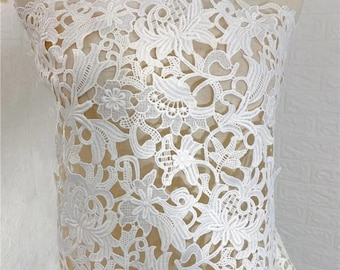 May include: Close-up of white floral lace fabric. The lace features a detailed pattern of flowers and leaves, creating a delicate and intricate design. The fabric is likely used for apparel or decorative purposes.
