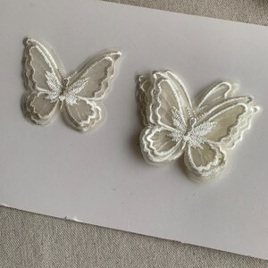 May include: Two white butterfly appliques with delicate embroidered details. The butterflies have a layered design, with a sheer, textured appearance. The wings are outlined with a fine, white thread, creating a detailed and elegant look.