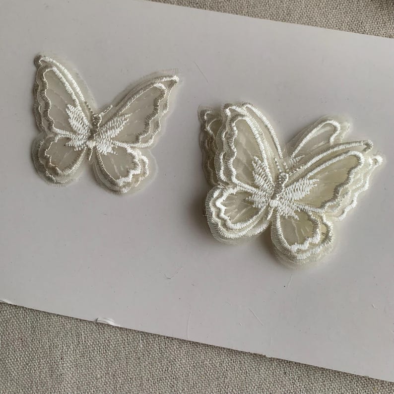May include: Two white butterfly appliques with delicate embroidered details. The butterflies have a layered design, with a sheer, textured appearance. The wings are outlined with a fine, white thread, creating a detailed and elegant look.