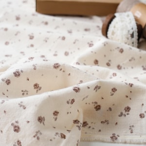 May include: Off-white fabric featuring a subtle floral design in brown tones. The fabric appears soft and has a slightly crumpled texture. A small brown box and a wooden spool with lace are in the background.
