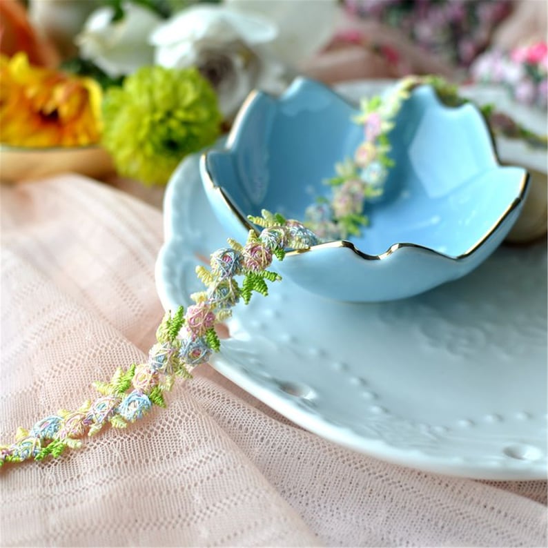 May include: A delicate floral lace trim with pastel pink, blue, and yellow flowers. The trim is displayed on a blue and white ceramic dish.