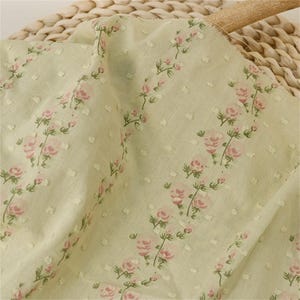 Pode incluir: Um tecido verde-sálvia claro com um padrão repetitivo de pequenos grupos de rosas cor-de-rosa dispostas em linhas verticais. O têxtil tem uma textura subtil com pequenos pontos brancos em relevo espalhados pela superfície do tecido.
