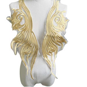 May include: Two gold embroidered appliques in the shape of stylized wings with a floral design. The appliques are on a white mesh background.
