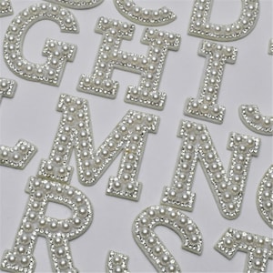 May include: A set of white pearl and rhinestone alphabet letters. The letters are all uppercase and are arranged in a random order.