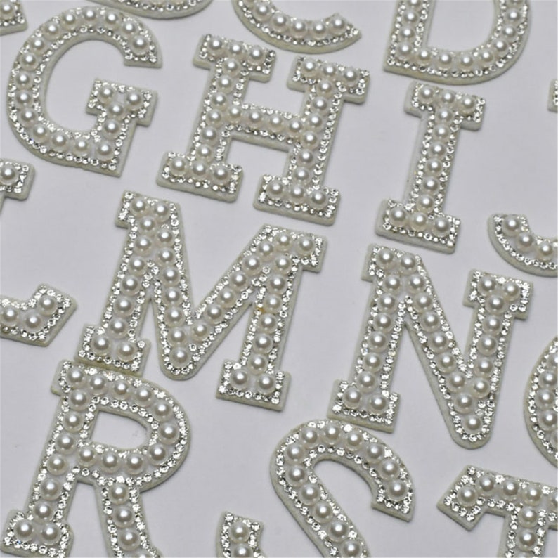 May include: A set of white pearl and rhinestone alphabet letters. The letters are all uppercase and are arranged in a random order.