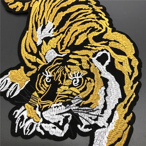 Può includere: Toppa ricamata a forma di tigre in nero, oro e bianco. La tigre è raffigurata in una posa dinamica, con cuciture dettagliate che creano un effetto strutturato. La toppa è probabilmente destinata all'applicazione su abbigliamento o accessori.