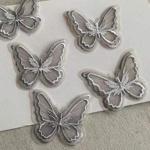 May include: Five grey butterfly appliques with detailed embroidery. The butterflies have a layered design with a translucent appearance. The wings are outlined in a lighter shade of grey.