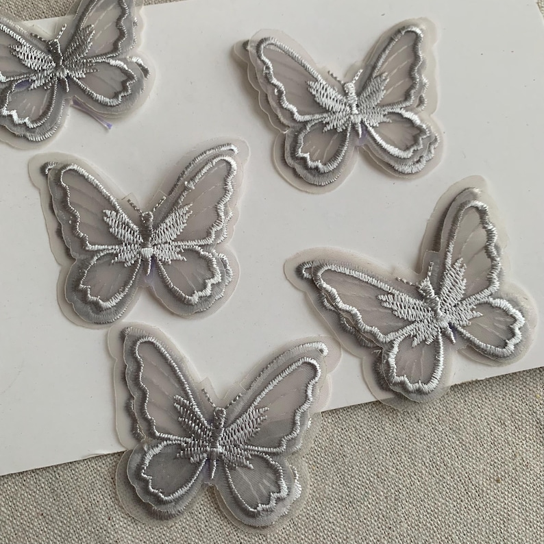 May include: Five grey butterfly appliques with detailed embroidery. The butterflies have a layered design with a translucent appearance. The wings are outlined in a lighter shade of grey.